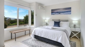 A bedroom with a large window, a bed with white bedding, a bench, and a painting on the wall.