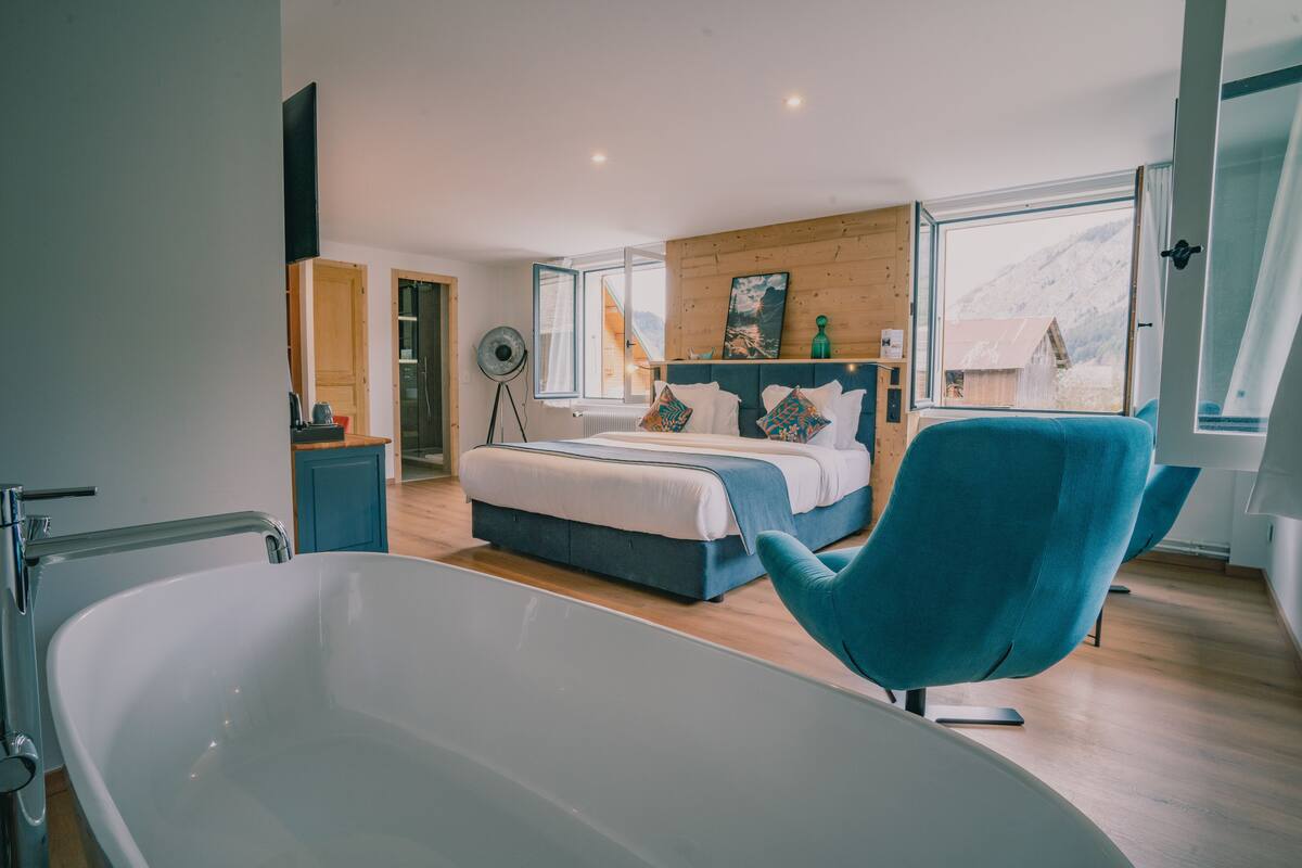 Ein modernes Schlafzimmer mit einer großzügigen Badewanne, einem Bett mit blauer Bettwäsche und Bergblick durch ein großes Fenster.