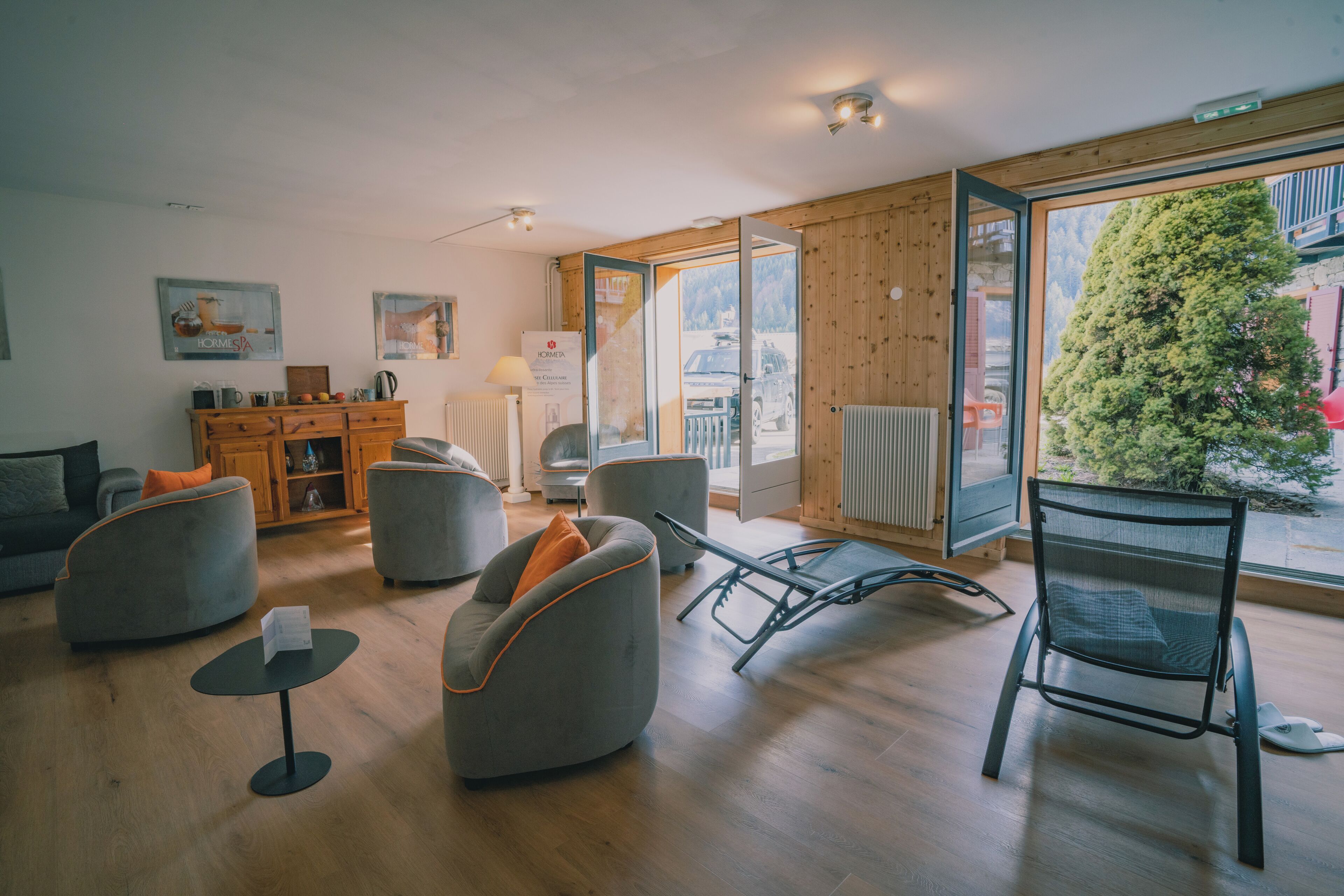 Un salon moderne avec un canapé, des fauteuils, une petite table et une chaise. On aperçoit un buffet en bois sur lequel sont posés divers objets. Une fenêtre donne sur l’extérieur.