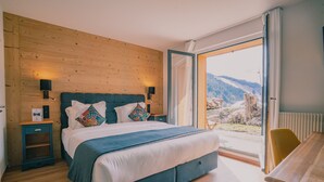 A bedroom with a large bed, wooden wall paneling, a nightstand, a desk, and a view of a mountainous landscape.