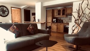 A modern living room with a grey sofa, two armchairs, a coffee table, and a woven ottoman. A wooden dining table and chairs are visible in the background.
