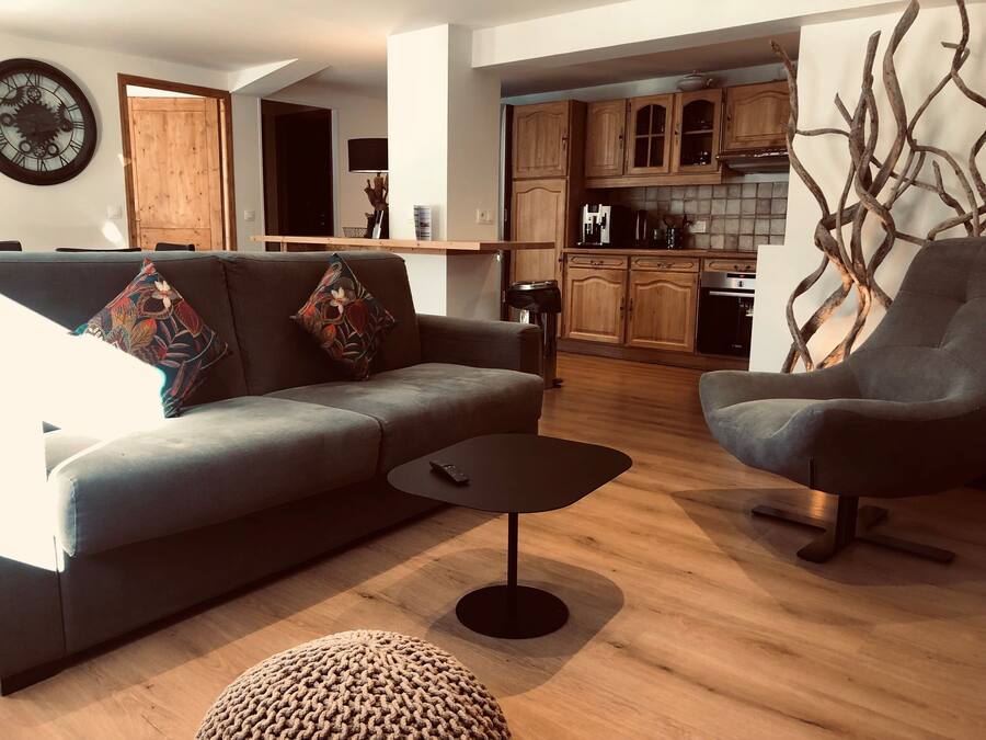 Un salon moderne avec un canapé gris, deux fauteuils, une table basse et un pouf en osier. On aperçoit une table et des chaises en bois à l’arrière-plan.