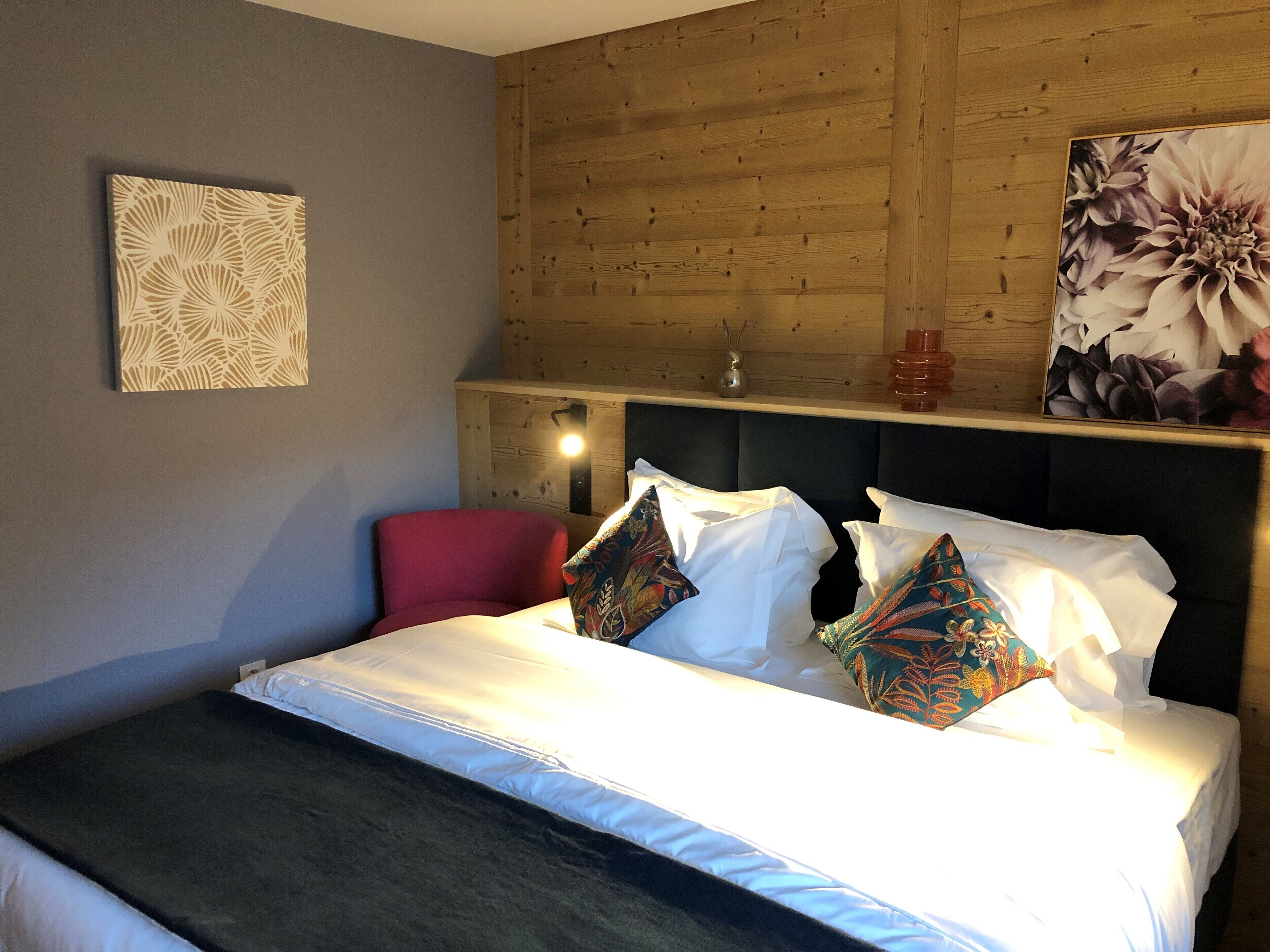 Une chambre à coucher avec un lit, des oreillers, un fauteuil rouge, une tête de lit en bois et un mur orné d’un tableau représentant des motifs floraux.