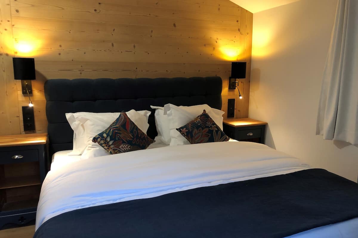 Ein ordentlich bezogenes Bett mit dunklem Kopfteil, zwei Nachttische mit Lampen und einer Holzbodenverkleidung.