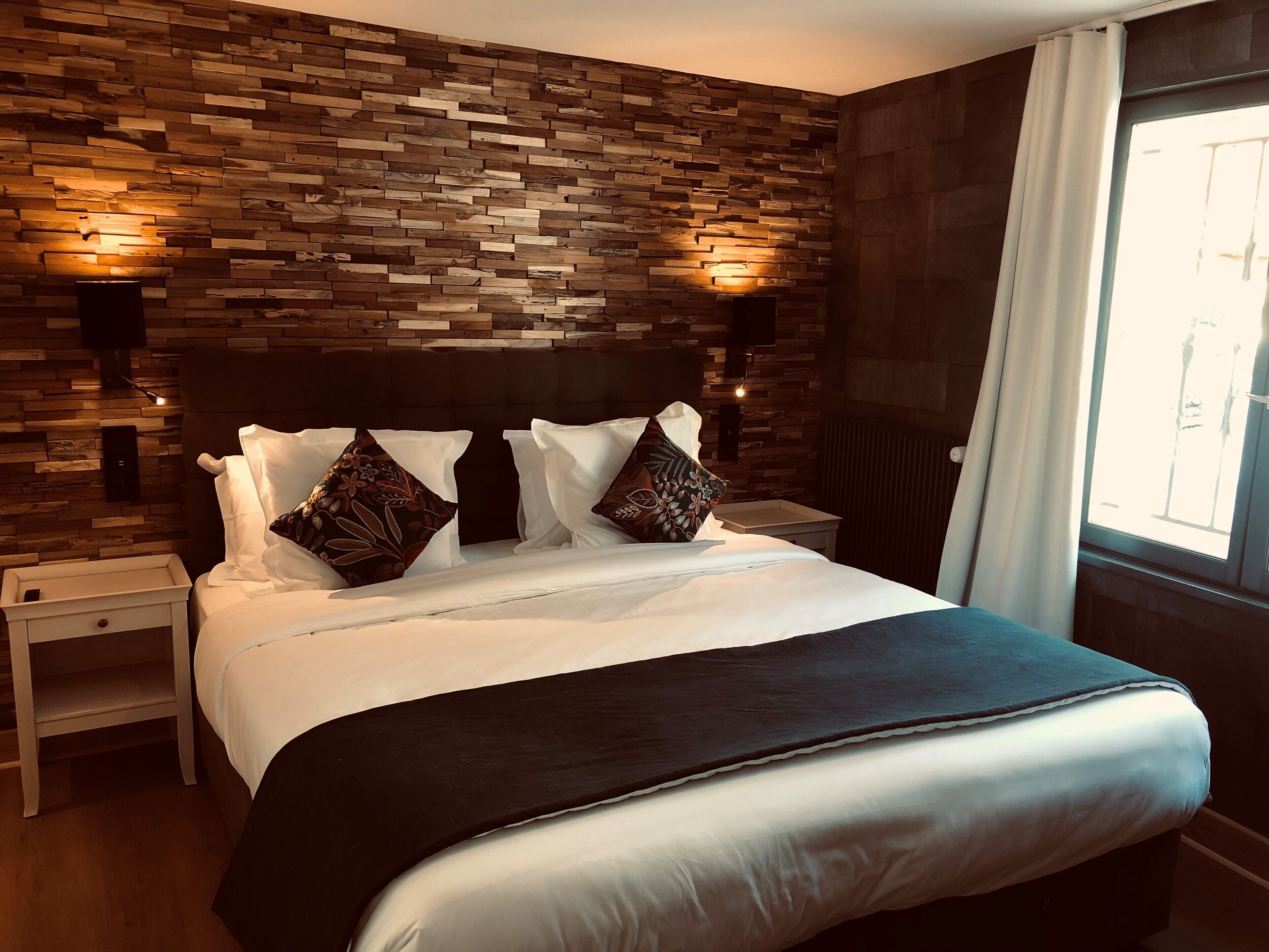 A hotel room with a wooden wall, a large bed with white bedding, two bedside tables, and a window with curtains.