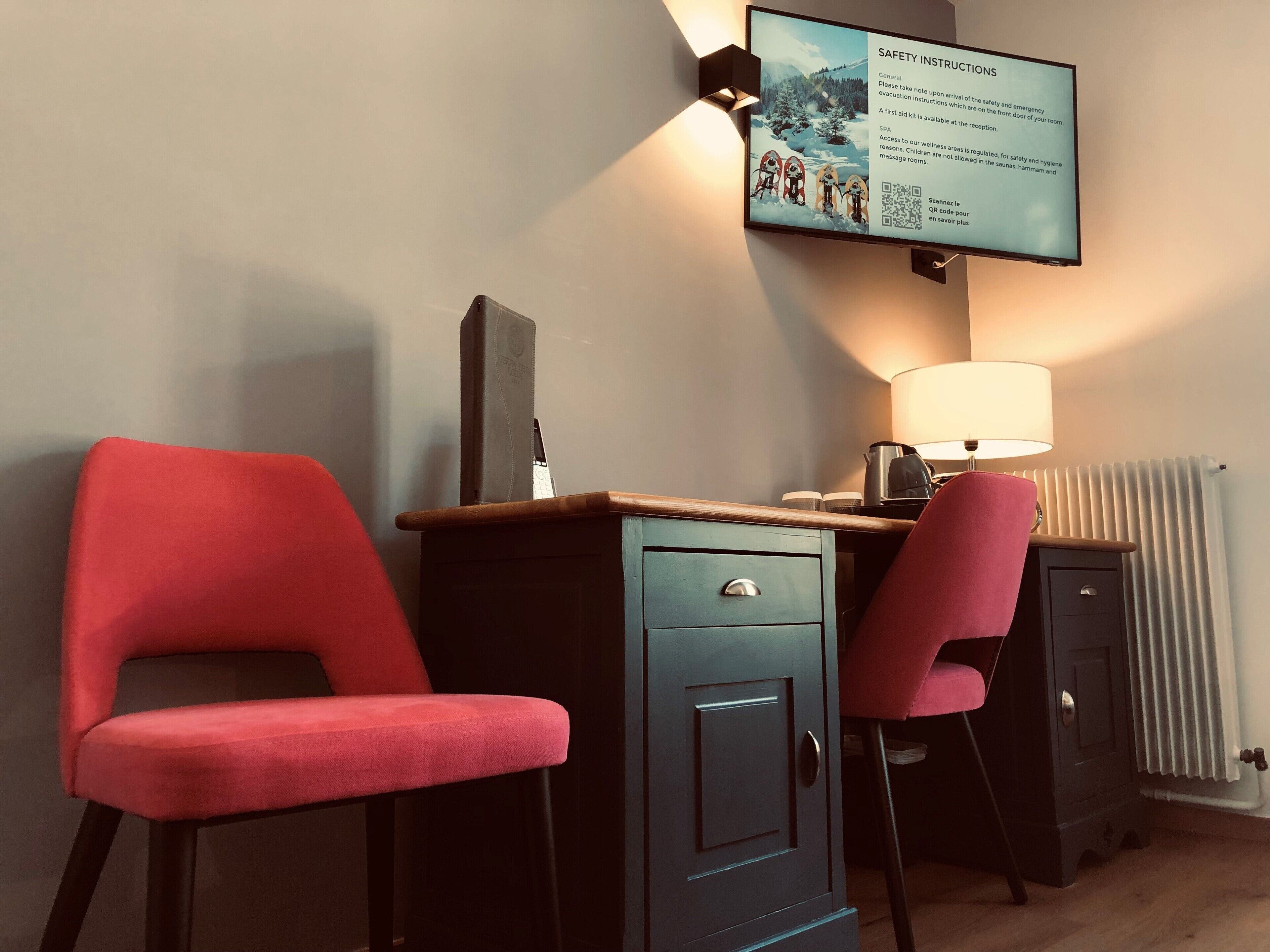 A hotel room with two red chairs, a desk with a lamp, a television displaying safety instructions, and a radiator.