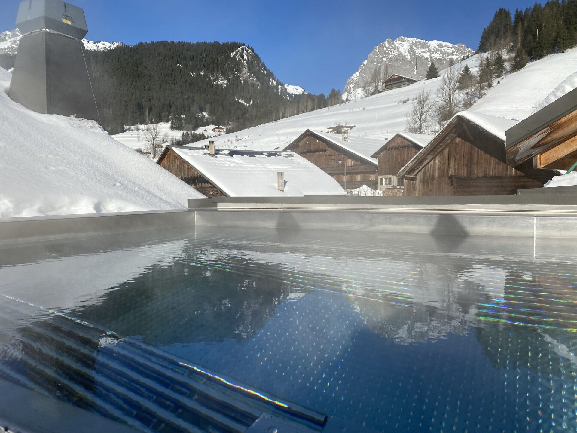 Una piscina cristalina con reflejos, rodeada de chalés de madera y montañas nevadas.