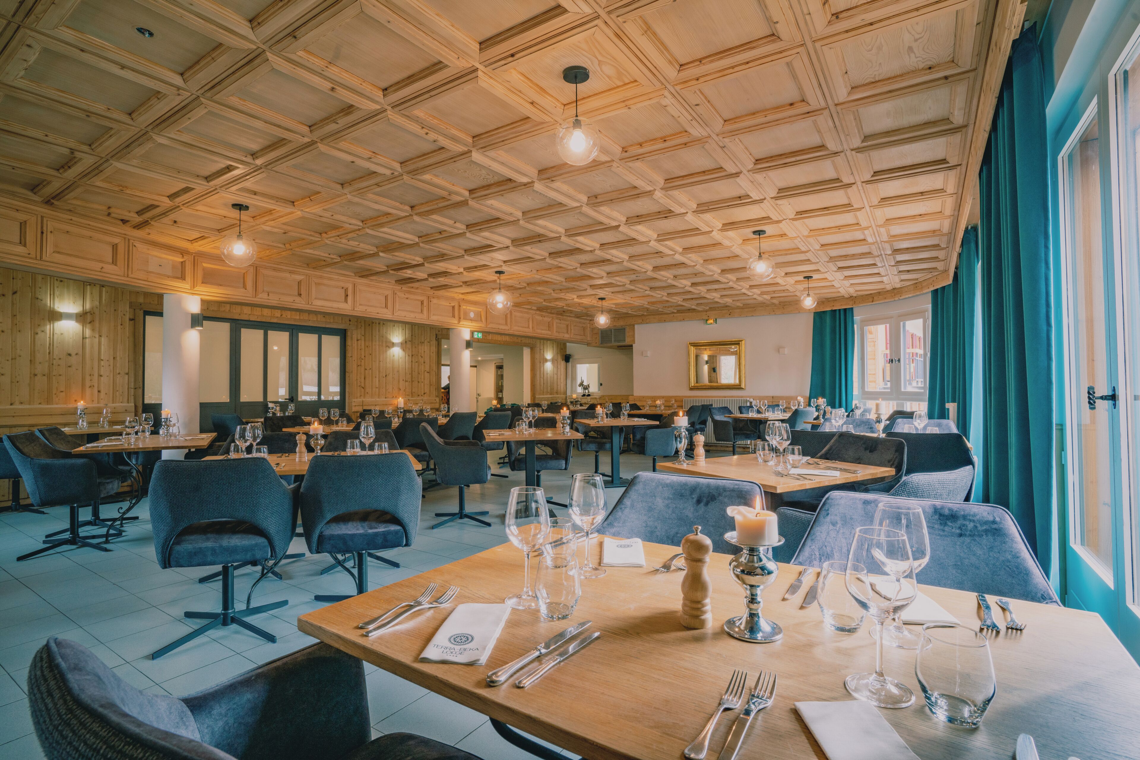 A spacious dining area with multiple tables and chairs, wooden ceiling, and large windows with curtains.