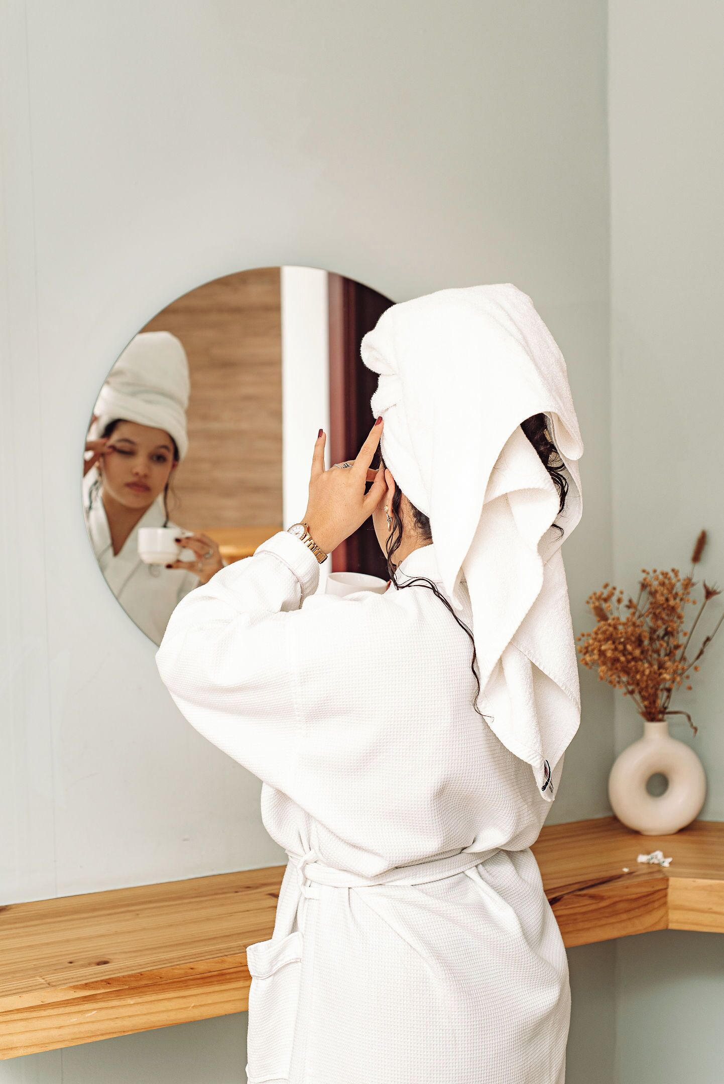 A person in a white robe standing in front of a mirror with a wooden shelf and a vase of dried flowers in the background.