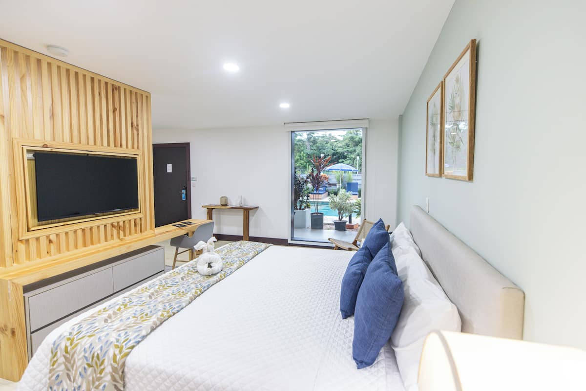 Une chambre dotée d’un grand lit, d’un téléviseur fixé sur un panneau en bois, d’un petit bureau et d’une porte-fenêtre coulissante donnant sur un balcon aménagé avec du mobilier d’extérieur.