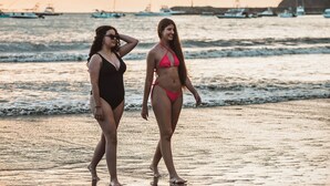 Twee personen wandelen over een strand, met boten op de achtergrond.