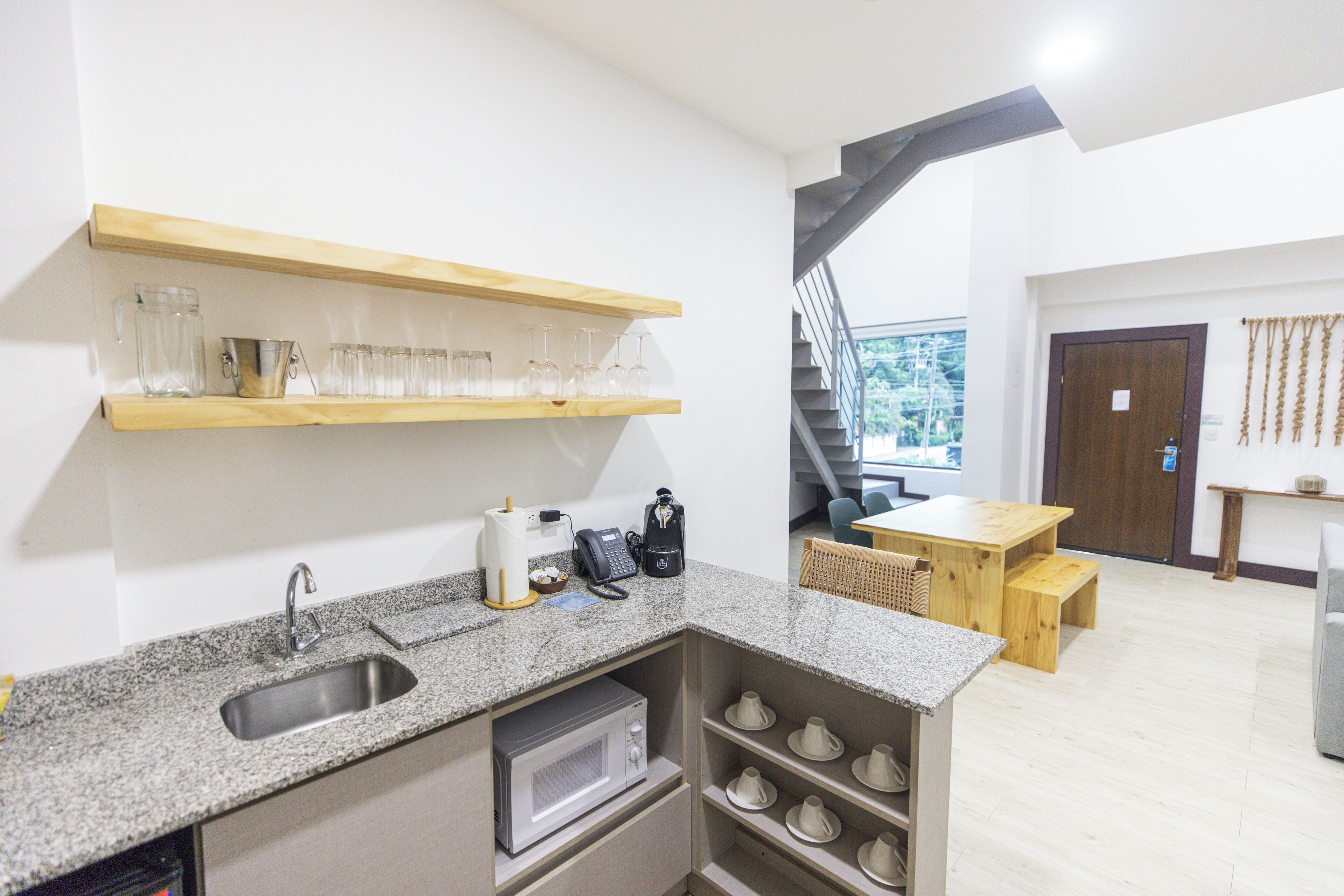 A compact kitchen with a granite countertop, a built-in microwave, and a coffee machine. There is a wooden shelf above the sink holding glassware. A staircase leads to a balcony with a view of greenery.