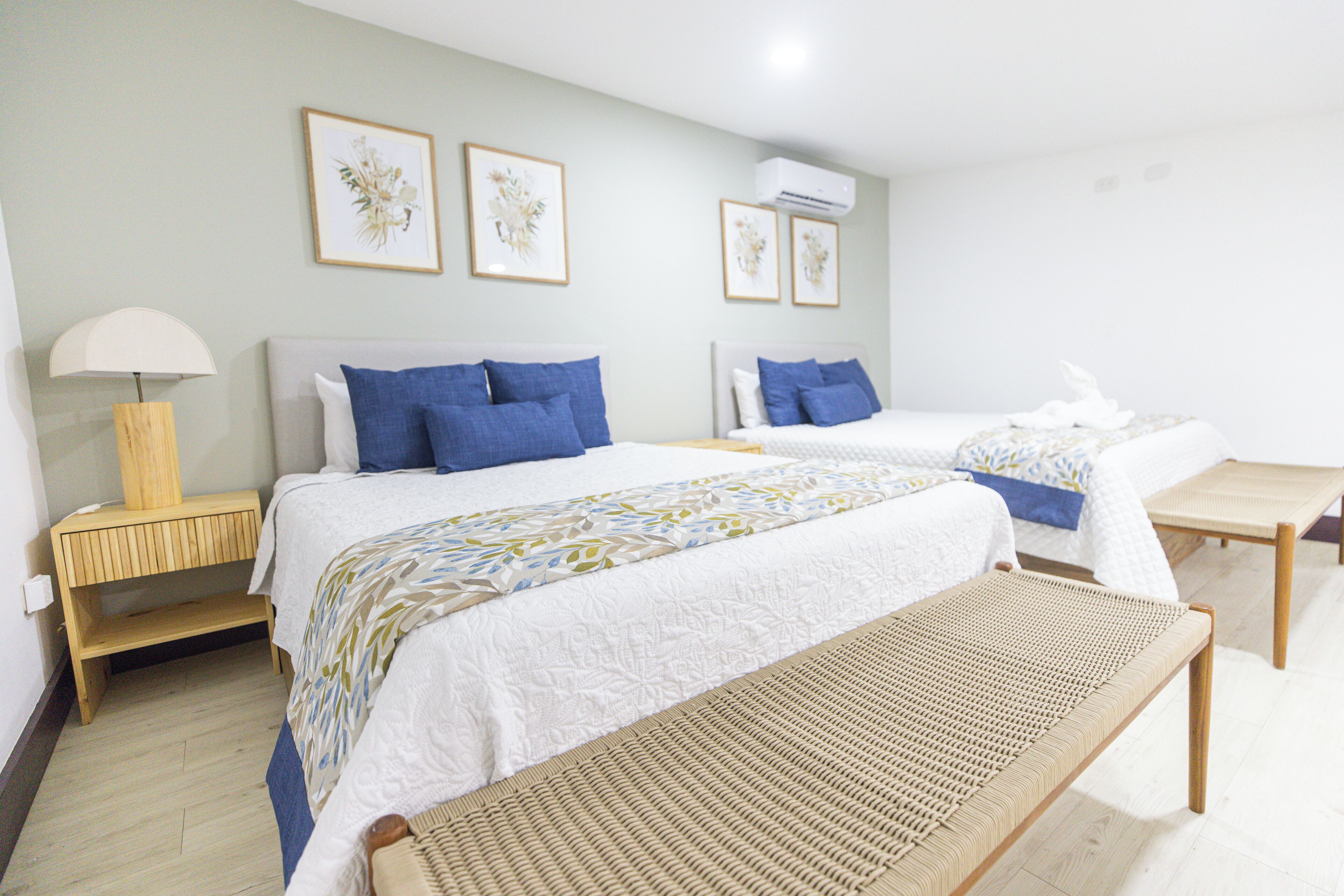 A hotel room with two beds, a bench, a nightstand, and wall-mounted air conditioning.