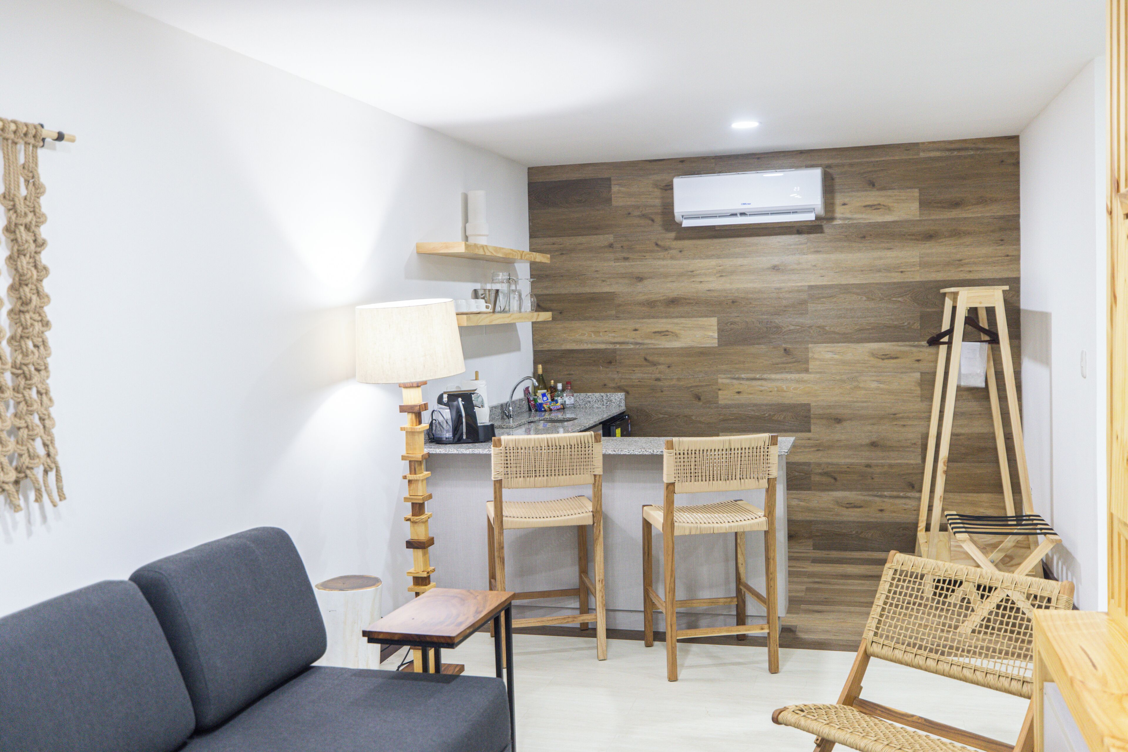 A modern room with a grey sofa, two wicker chairs, a small wooden table, a lamp, a kitchen area with a counter and chairs, and a wooden wall with a mounted air conditioner.