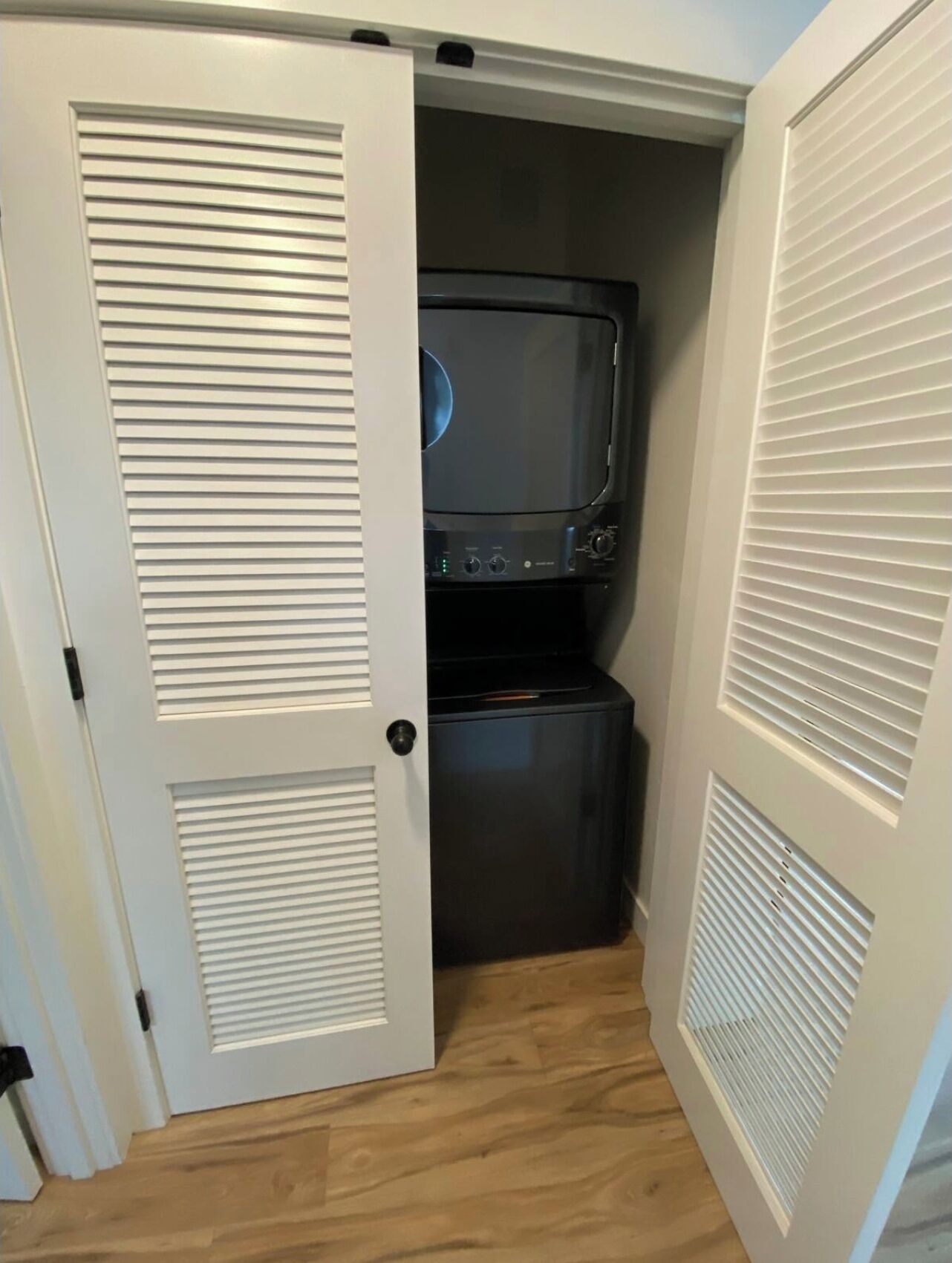 A compact laundry room with a washer and dryer, white doors with louvered panels, and a wooden floor.