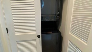 A compact laundry room with a washer and dryer, white doors with louvered panels, and a wooden floor. - Base Camp for Bend Explorers (Bend)