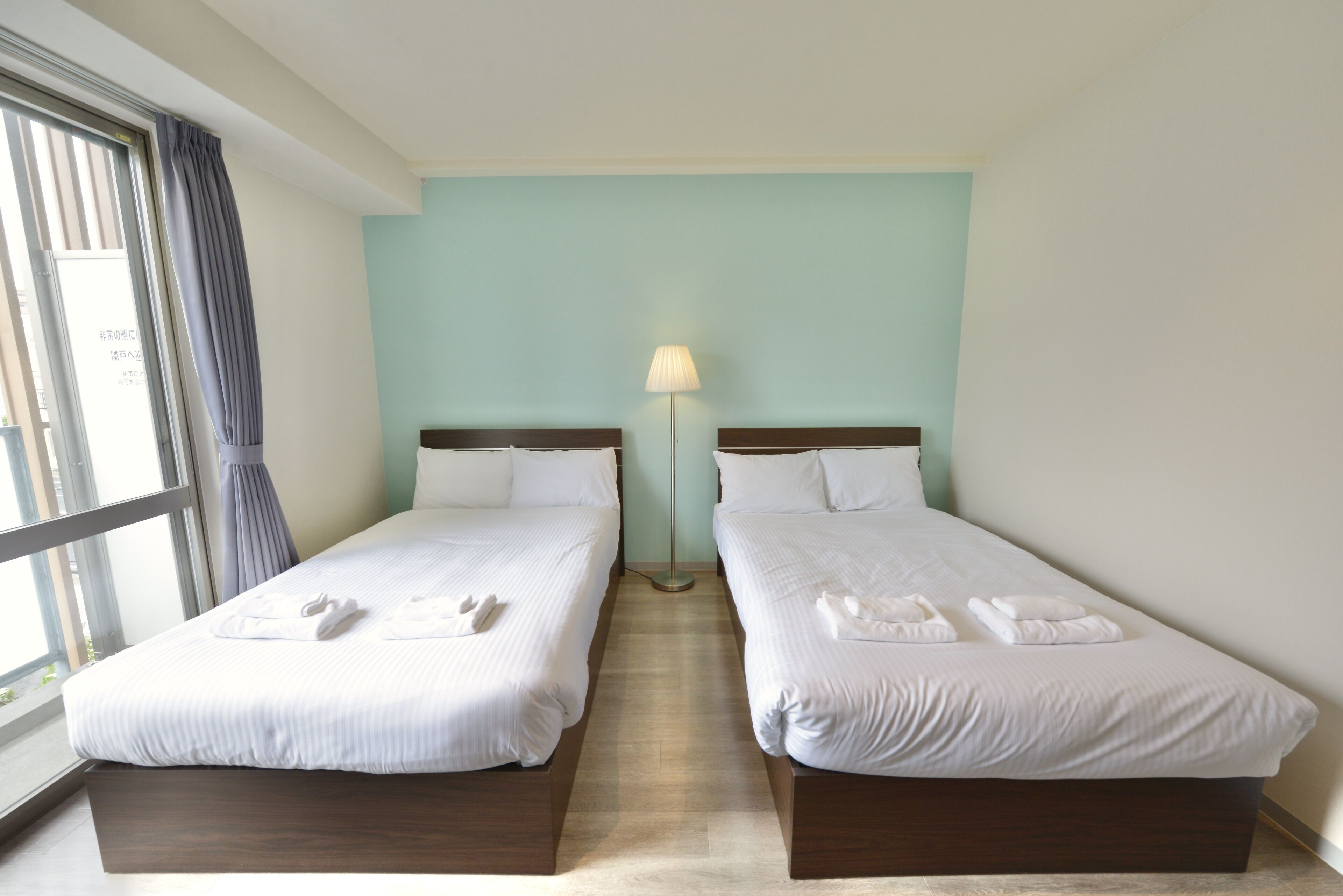 A hotel room with two beds, a window with curtains, a standing lamp, and a view of the outside.