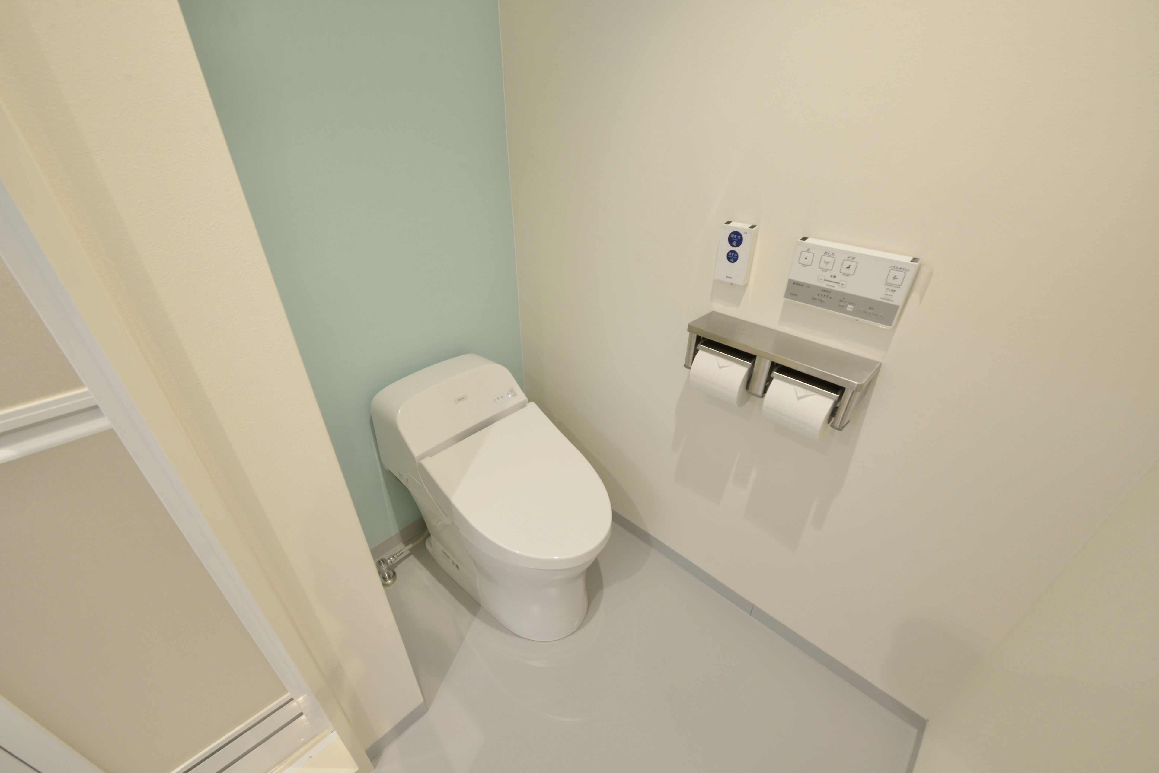 A small, modern bathroom with a white toilet, a wall-mounted paper towel dispenser, and a control panel.