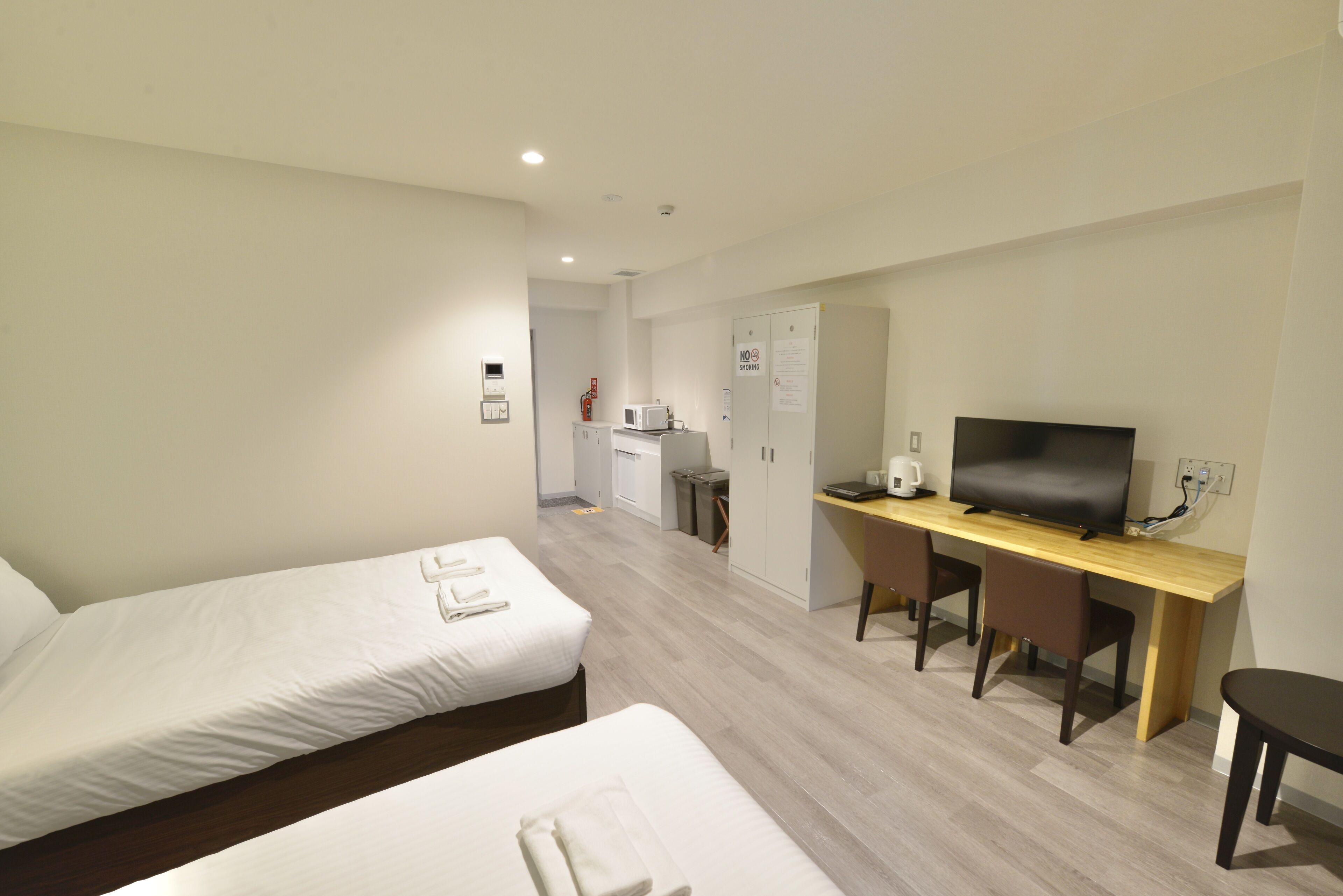 Une chambre d’hôtel avec deux lits, un bureau avec une télévision et, au fond, une petite cuisine.