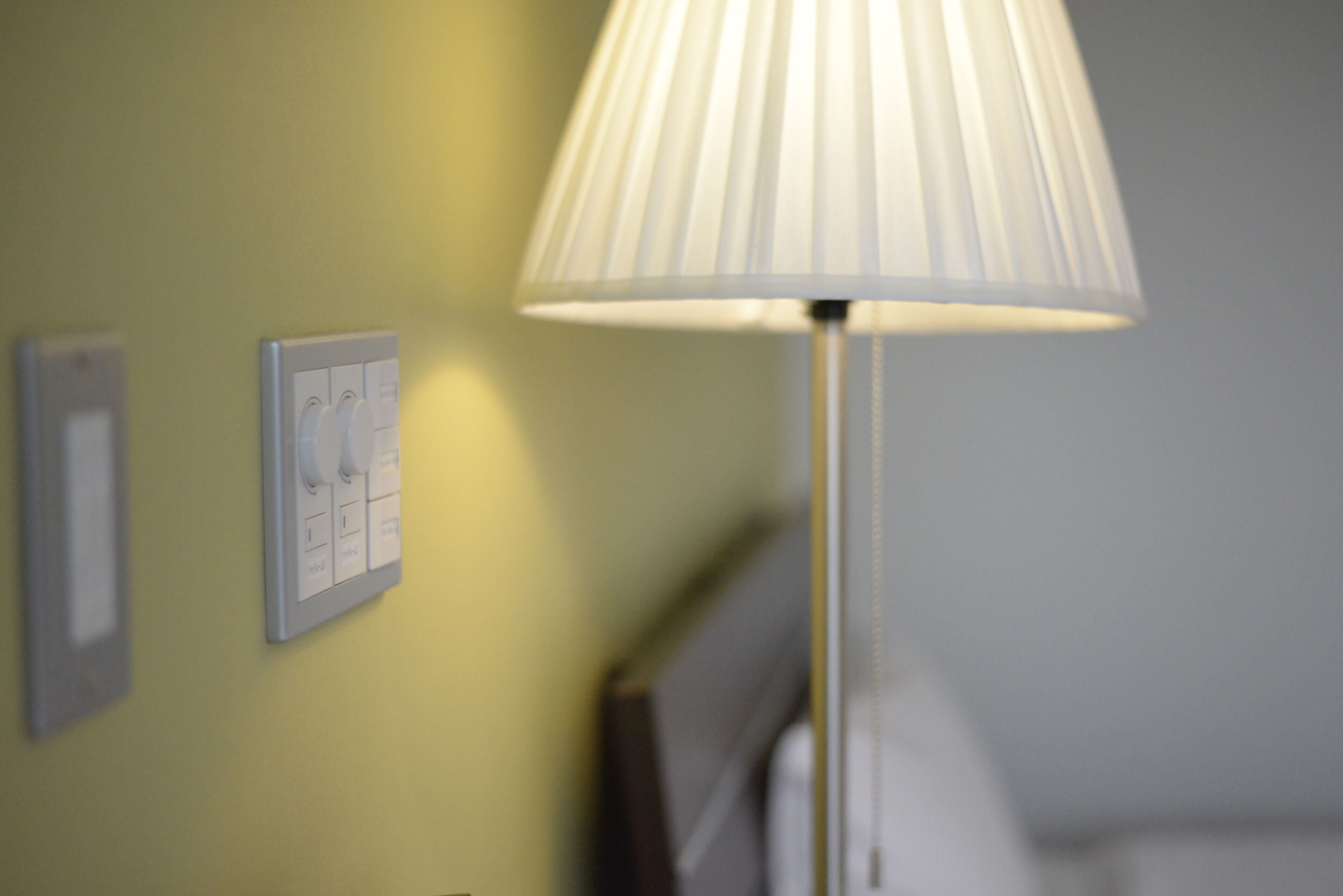 A lamp with a white shade, a wall switch, and a framed picture on a yellow wall.