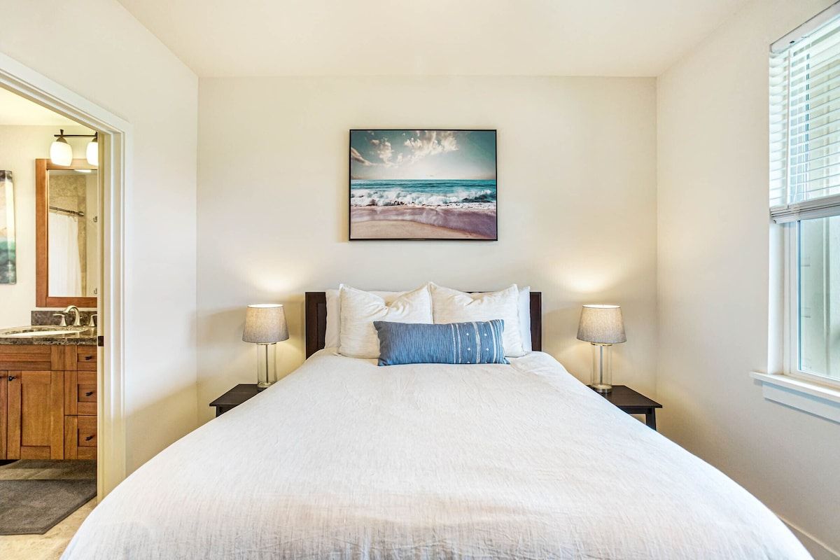 A bedroom with a bed, two bedside lamps, a painting of a beach scene, and a window with blinds.