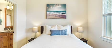 A bedroom with a bed, two bedside lamps, a painting of a beach scene, and a window with blinds.