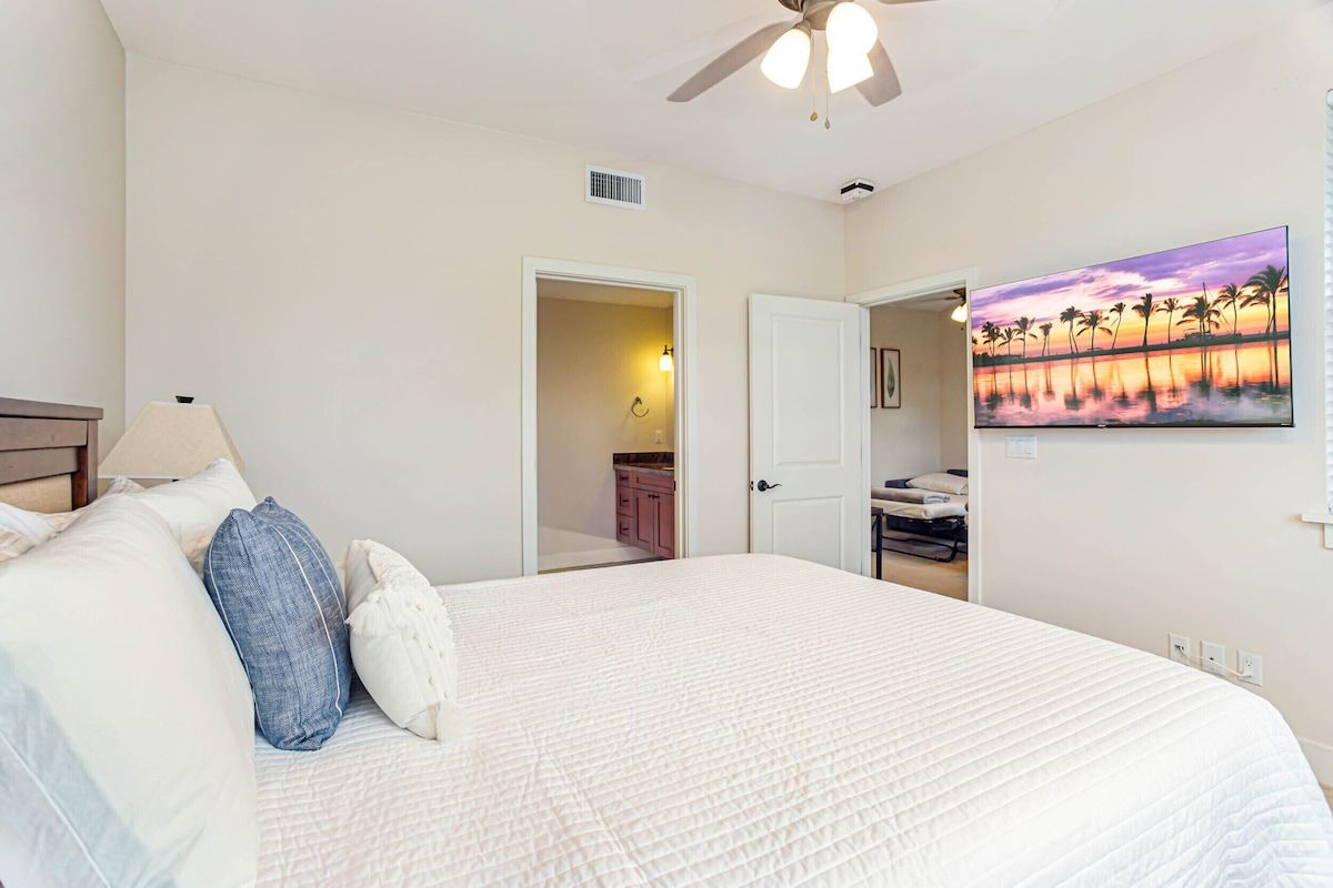 Une chambre à coucher avec un lit, des oreillers, un ventilateur de plafond, un tableau et une salle de bain visible à travers une porte ouverte.