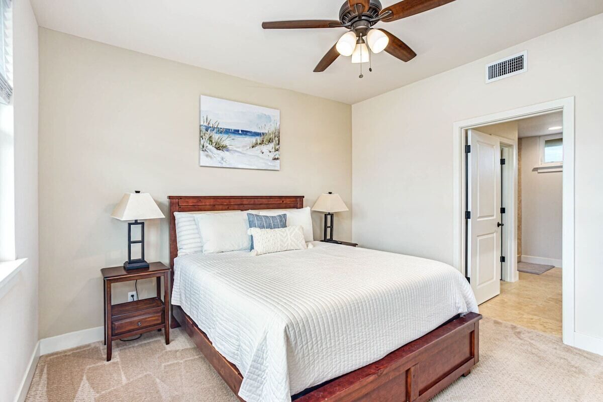 A bedroom with a wooden bed, two bedside tables with lamps, a ceiling fan, and a door leading to another room.