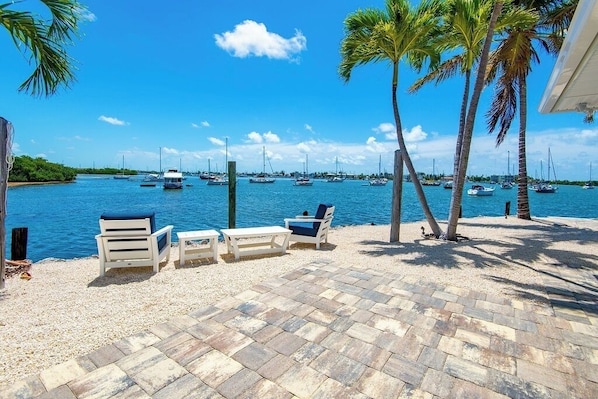 A beachfront patio with lounge chairs and a table, overlooking a marina with boats.