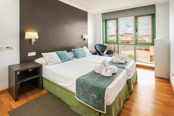 Habitación de hotel con dos camas, una mesita de noche, una silla y un ventanal con vistas.