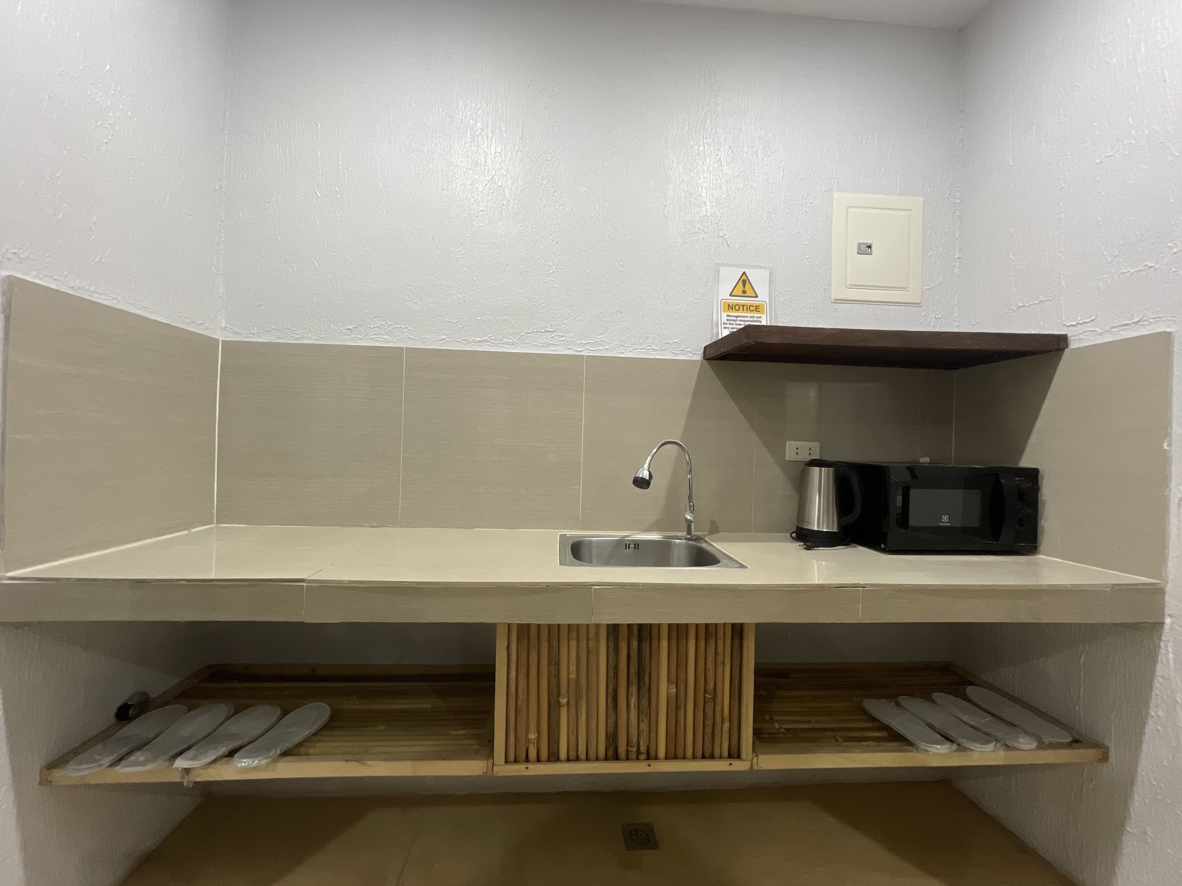 Una pequeña cocina equipada con un fregadero, cafetera y microondas. Hay un cartel en la pared y un estante con vajilla debajo de la encimera.