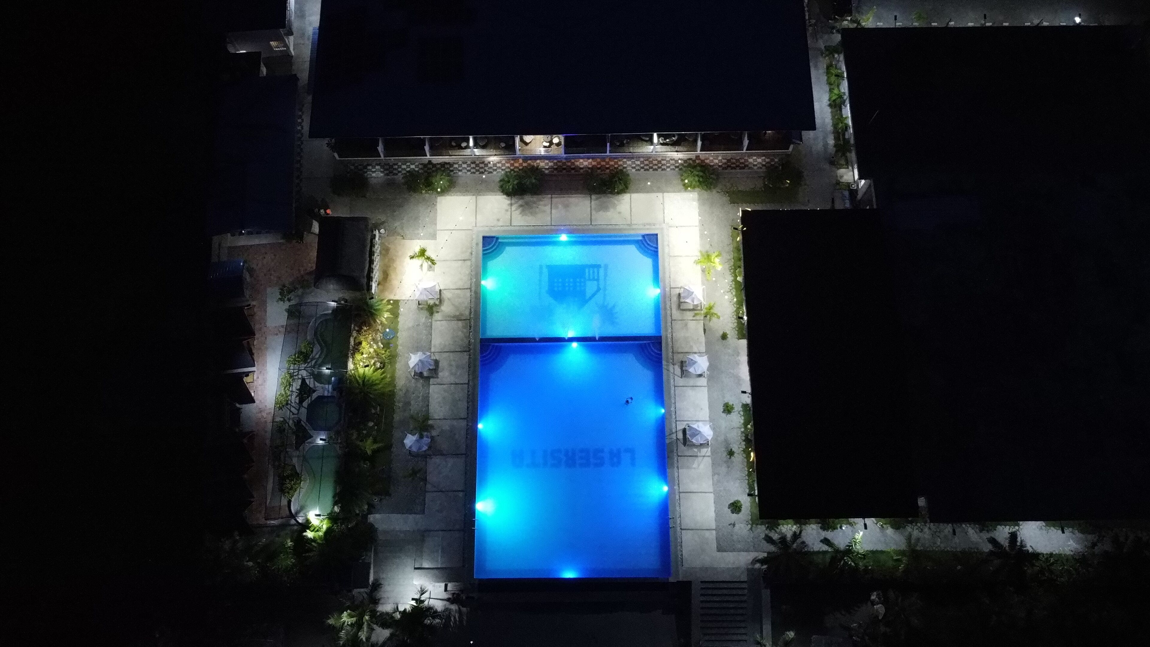 Vista aérea de una piscina con sillones de descanso y sombrillas alrededor, rodeado de vegetación y edificios.