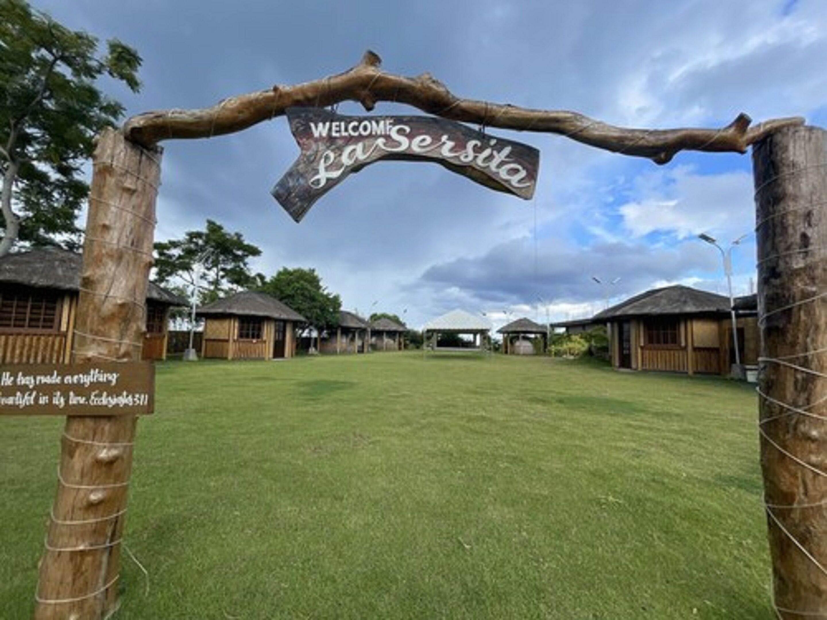 A rustic welcome sign with a wooden archway, a grassy lawn, and traditional huts in the background.