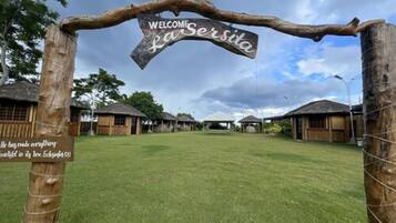 A rustic welcome sign with a wooden archway, a grassy lawn, and traditional huts in the background.