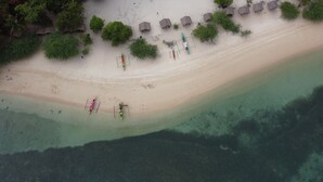 Une vue aérienne d’une plage aux eaux cristallines, avec plusieurs huttes aux toits de chaume et quelques personnes près de l’eau.