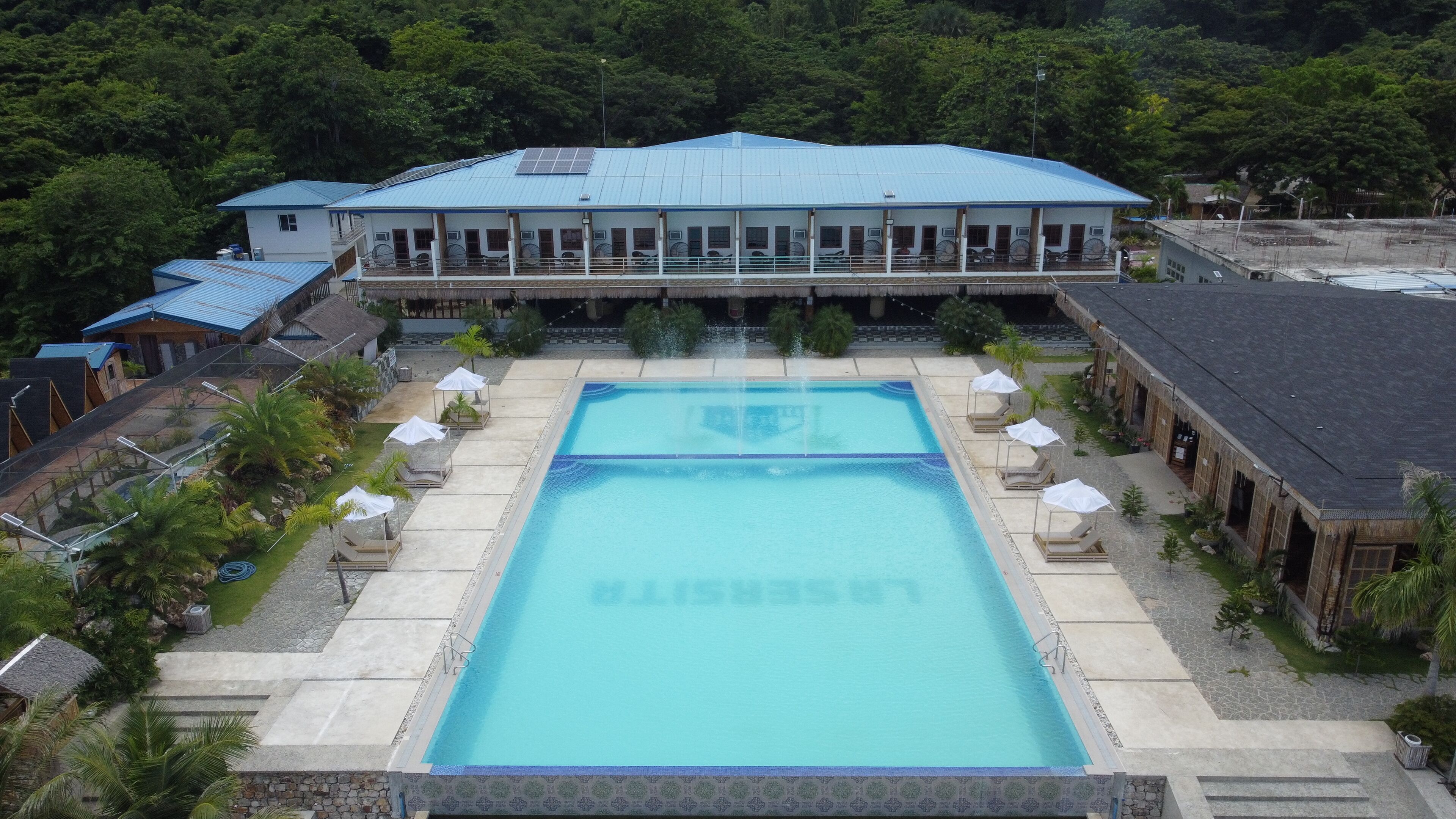 Una piscina rectangular grande con borde azul, rodeada de sillas de descanso y sombrillas, y un edificio al fondo.