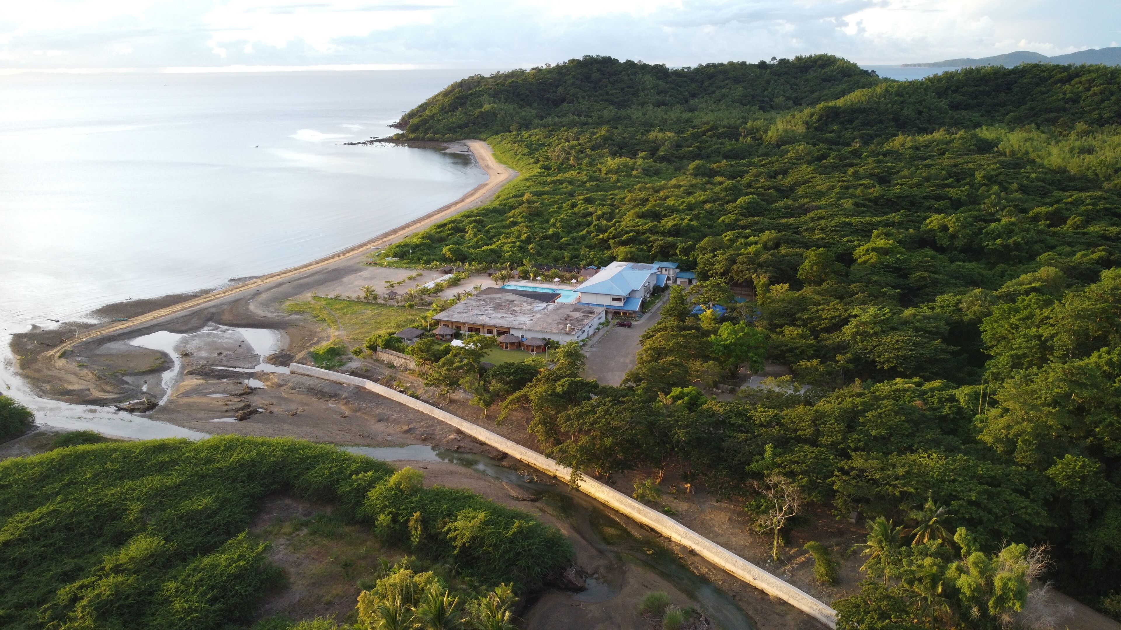 An aerial view of a coastal property with a beach, a swimming pool, and a large building with a blue roof.