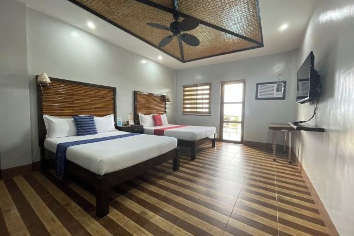 Habitación de hotel con dos camas, un ventilador de techo, un televisor montado en la pared y una ventana con persianas.