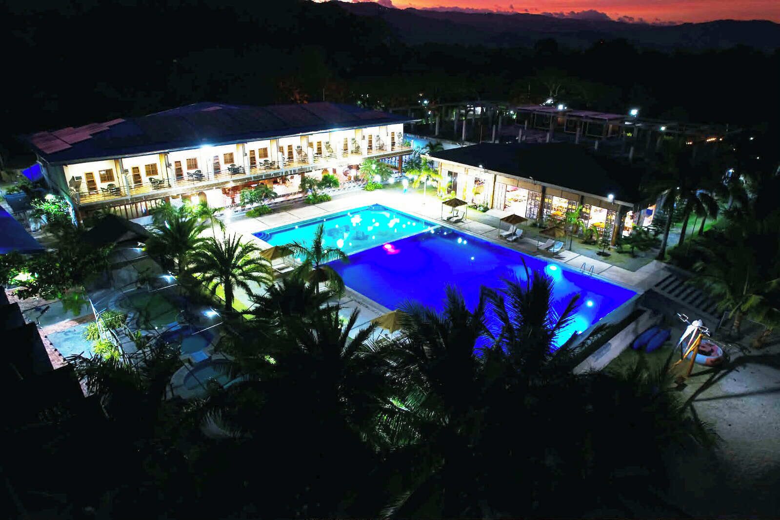 Um edifício grande com piscina, rodeado por palmeiras e iluminado à noite.
