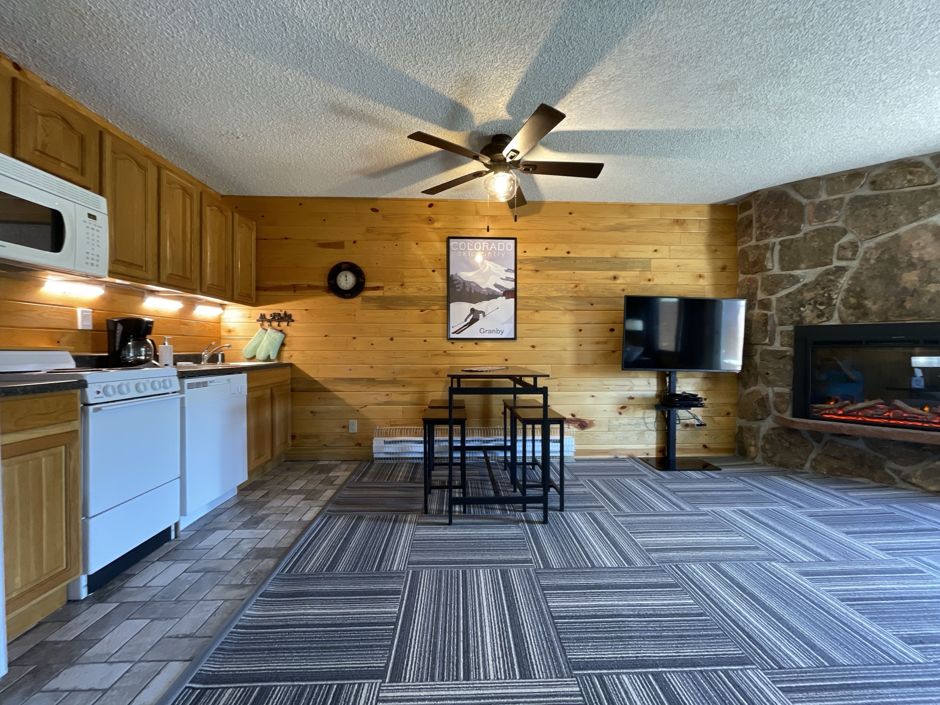 A kitchen with wooden cabinets, a microwave, a stove, a coffee maker, a clock, a television, and a stone fireplace.