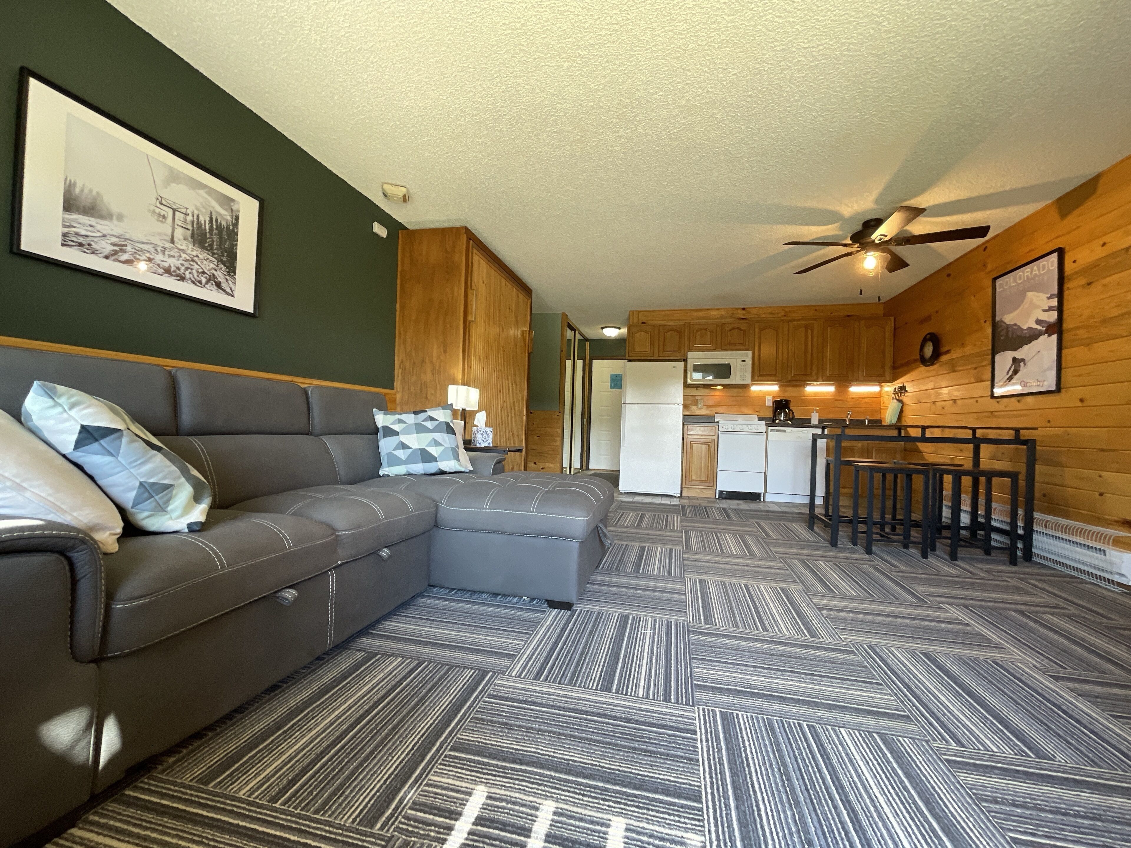 A modern living room with a sectional sofa, a kitchen area with wooden cabinets, and a ceiling fan.