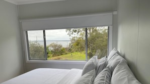 A bedroom with a large window, a bed with white linens, and a view of greenery and water outside.