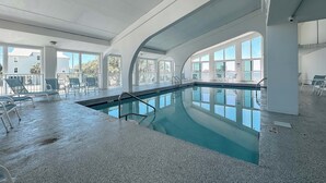 Una piscina interior con vistas despejadas a través de amplios ventanales, una zona de descanso junto a la piscina y un techo con vigas estructurales visibles.