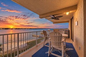 A balcony with a view of the sunset over the water, a table with chairs, and a ceiling fan.