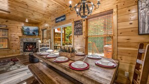 L’intérieur d’un chalet en bois présente une table à manger dressée pour quatre personnes, une cheminée et un panneau invitant les convives à s’asseoir.