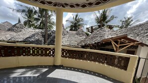 A covered outdoor area with a tiled floor, a decorative railing, and thatched-roof structures in the background. - Beautiful 3 Bedroom Villa in Diani Beach-sleeps 8 (Diani)