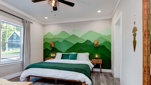 Slaapkamer met een bed, houten hoofdbord, een plafondventilator, een muurschildering, bedlampen en een raam met gordijnen.