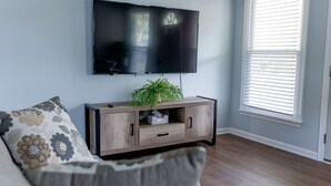 Uma sala de estar com televisão de tela plana na parede, um armário de madeira com uma planta e uma janela com persianas.