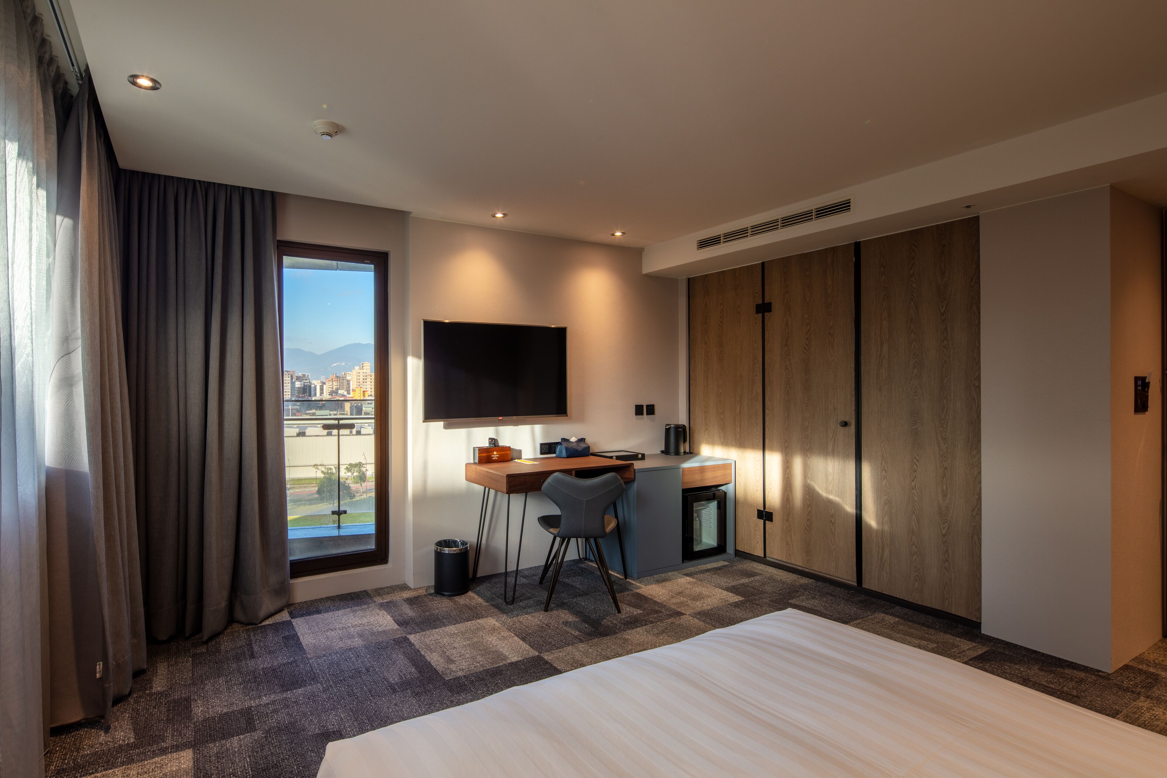 A hotel room with a large bed, a desk with a chair, a TV mounted on the wall, and a wardrobe.