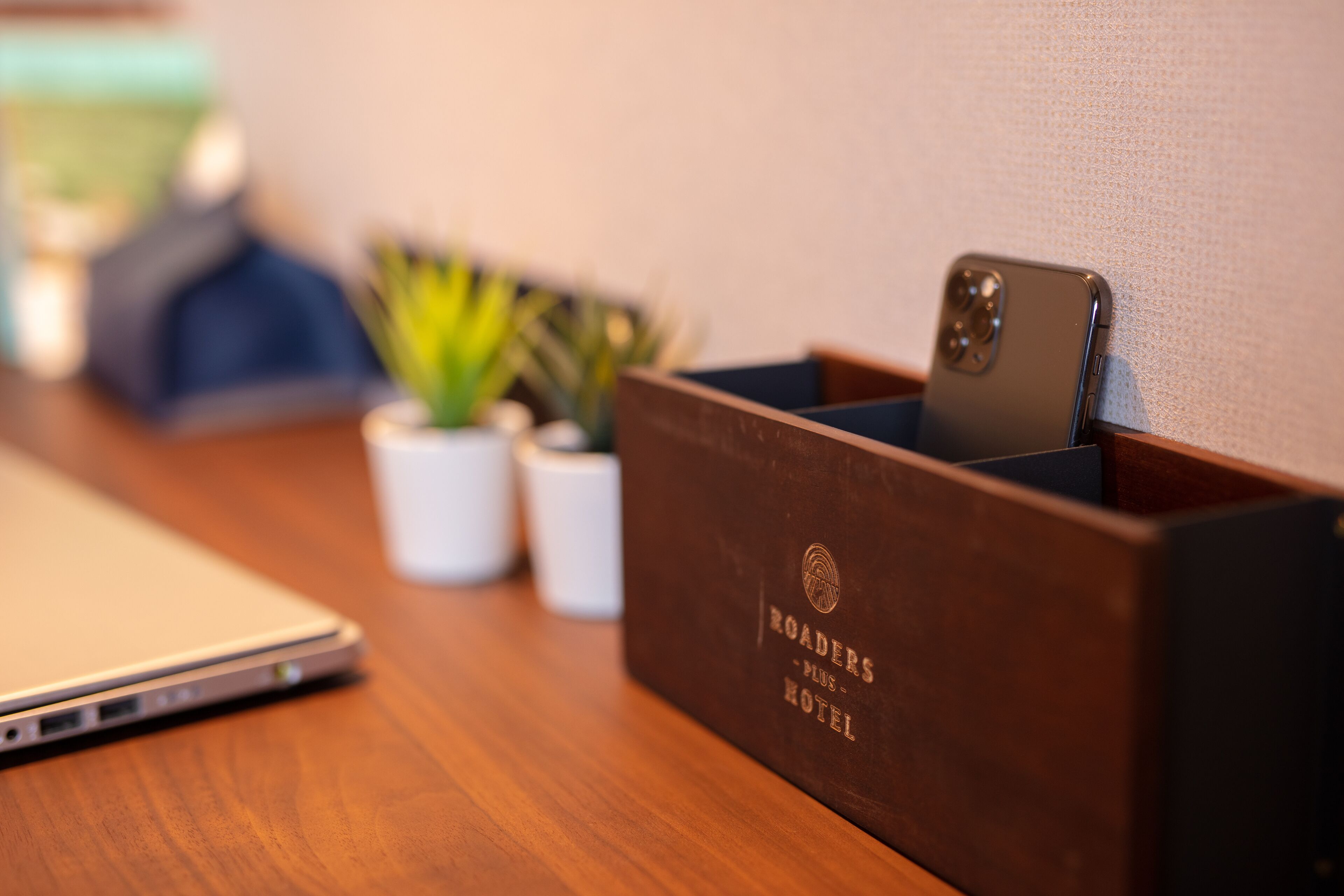 Un bureau en bois avec un ordinateur portable, un smartphone dans un support en bois, et deux petites plantes en pot.